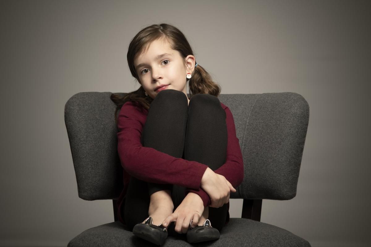 a girl in a burgundy raglan sits on a chair with her hands on her knees