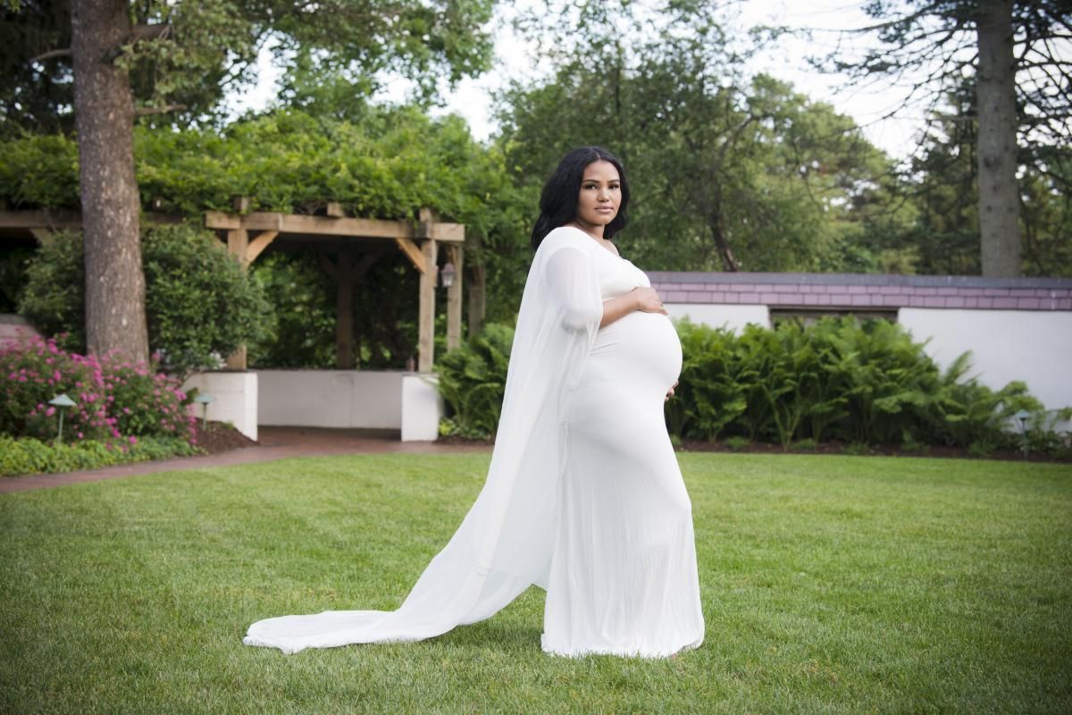 a pregnant woman stands on the lawn in a white dress gently stroking her belly