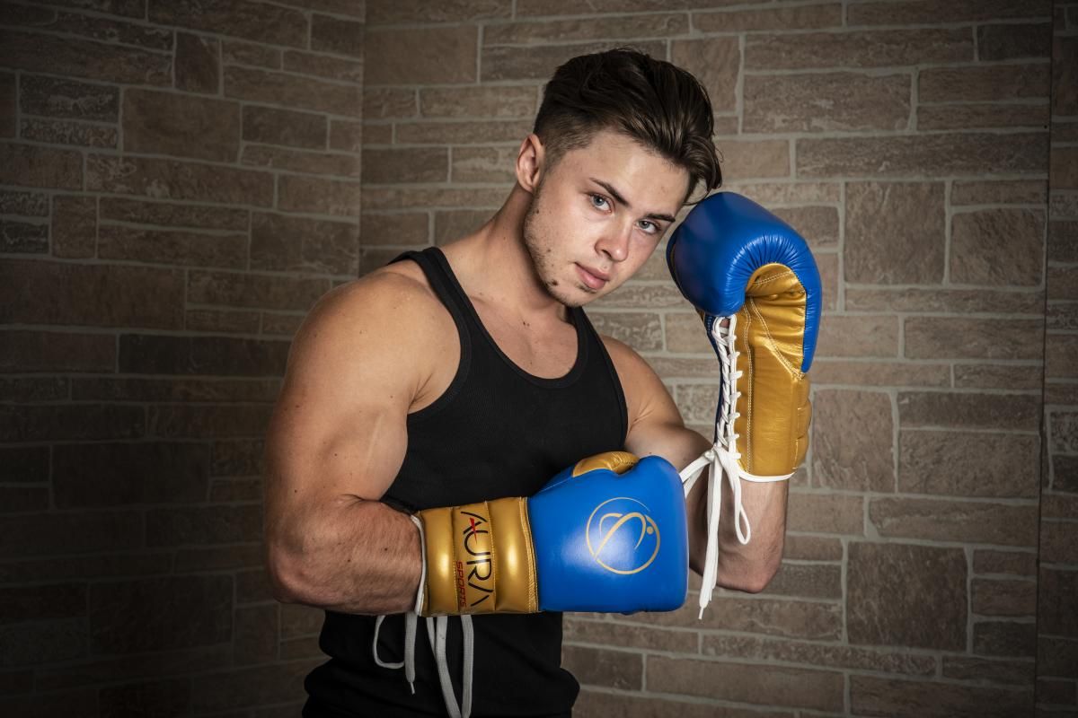a guy with dark hair and a black t-shirt in blue and gold boxing gloves aura sports poses in defense