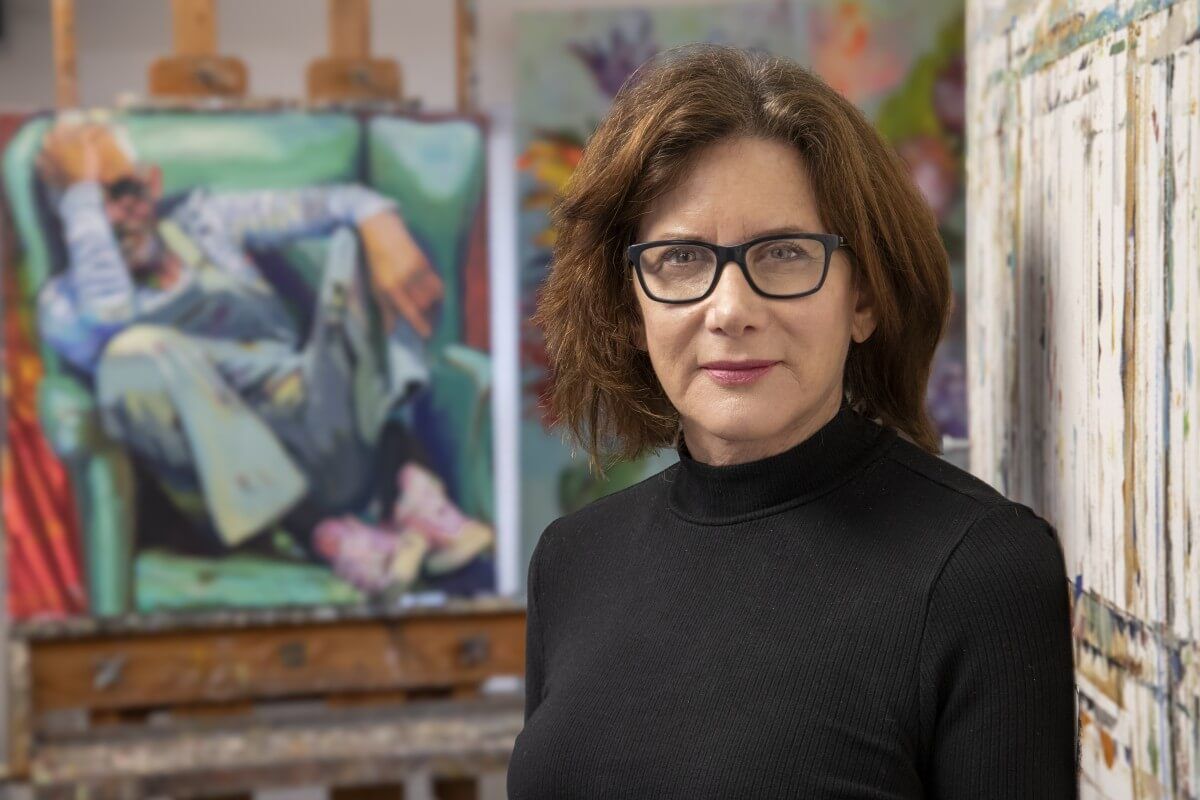 Portrait of a female artist with brown hair and black glasses, wearing a black turtleneck, standing in an art studio with a colorful painting on an easel in the background
