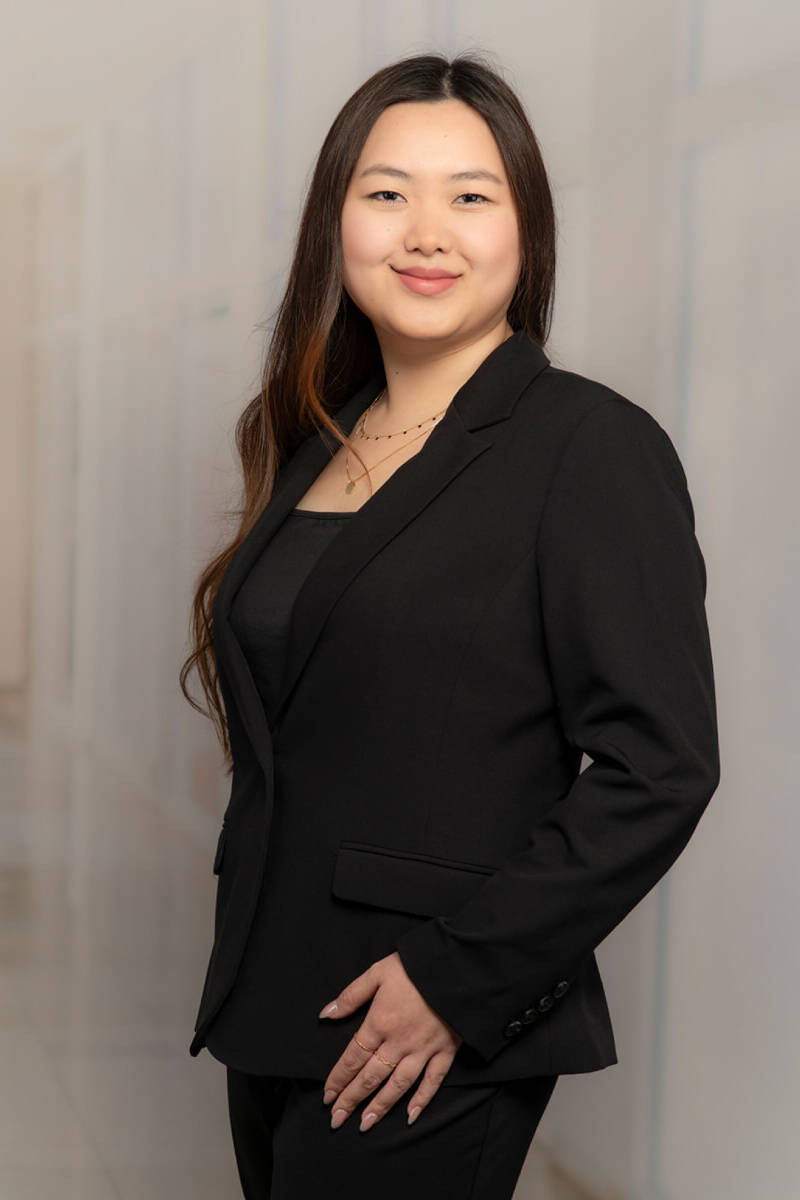 A brown-eyed, dark-haired girl in a black graduation gown with a square master's cap. Behind her is a gray background.