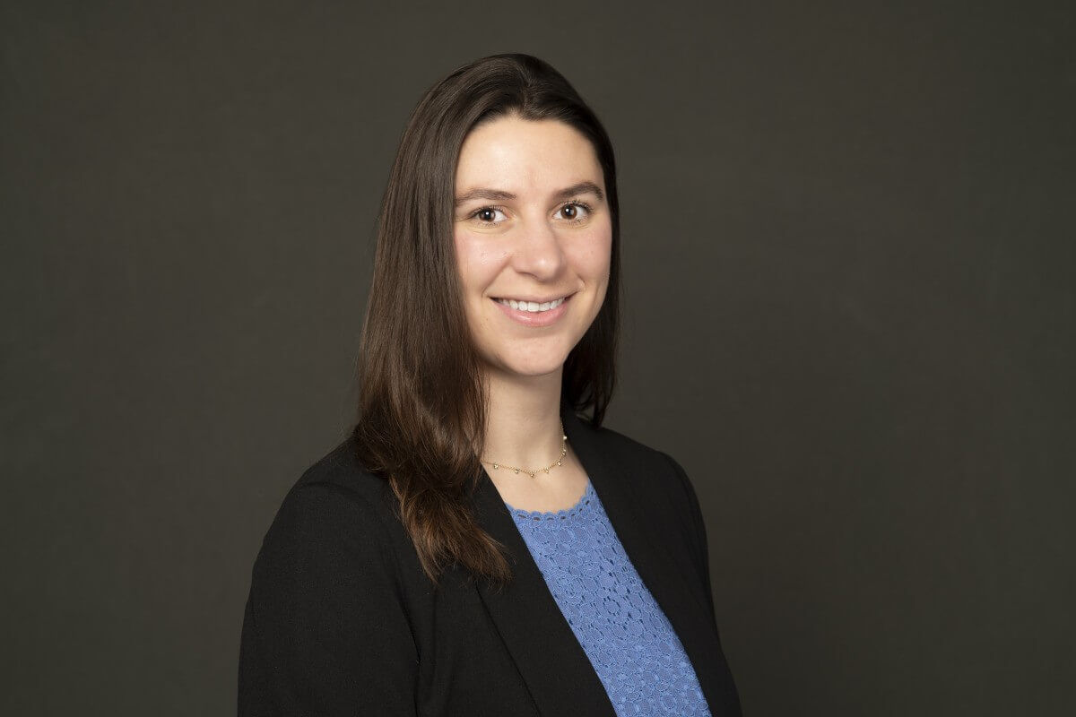 Professional headshot of a smiling woman with long brown hair wearing a black blazer and blue lace top against a dark grey background