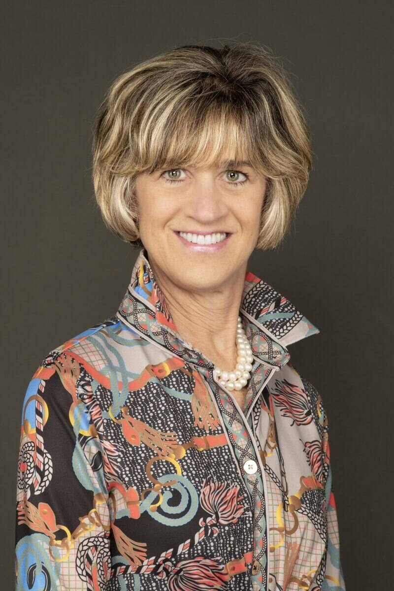Professional headshot of a smiling woman with blonde highlighted hair, wearing a colorful patterned blouse and a pearl necklace against a dark grey background
