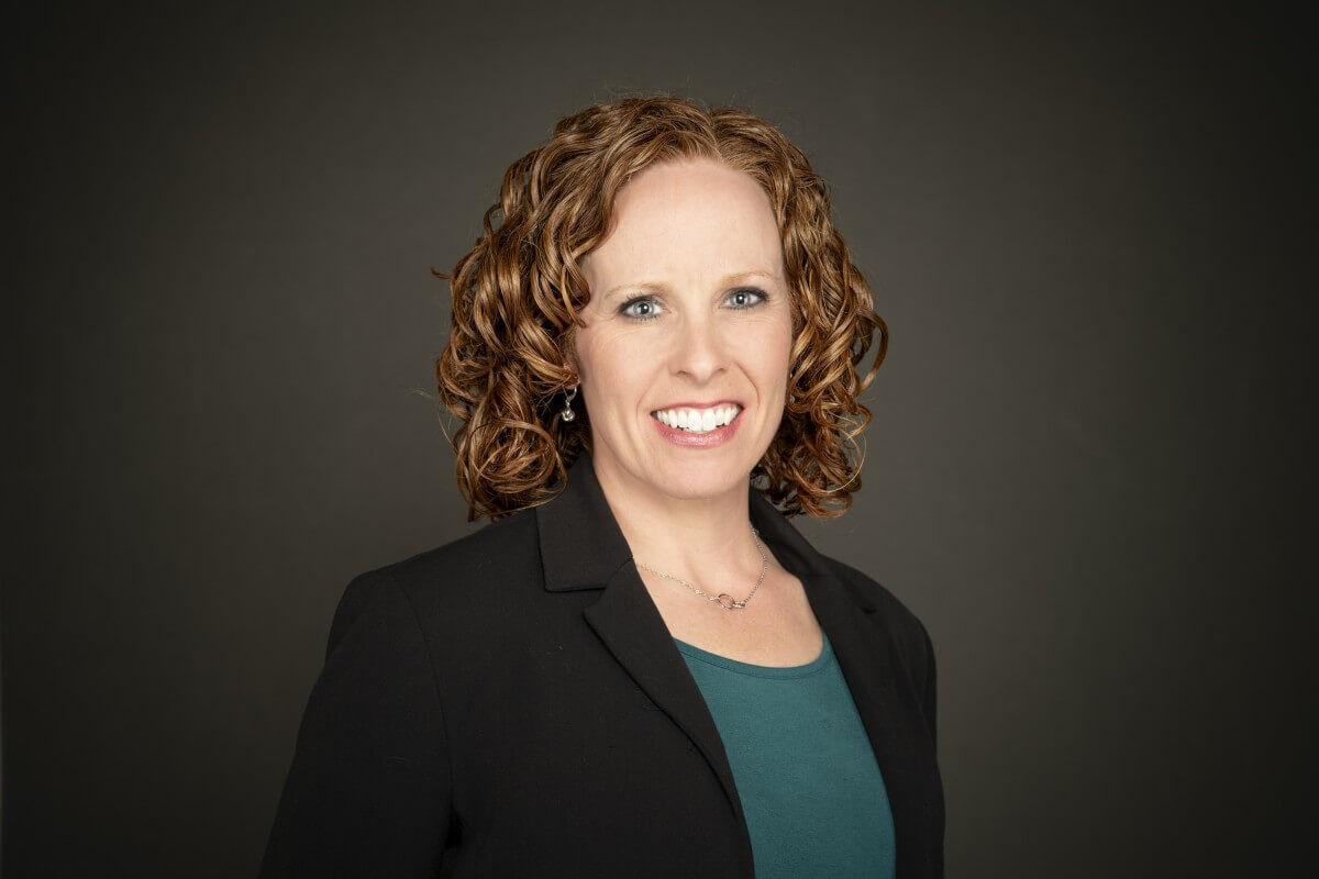 Professional headshot of a smiling woman with curly auburn hair, wearing a black blazer and teal top against a dark studio background