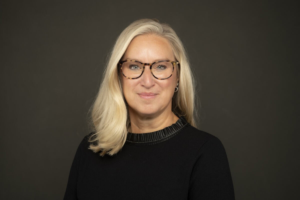 Professional portrait of a woman with blonde hair and tortoiseshell glasses, wearing a black top with a pleated collar against a dark grey background