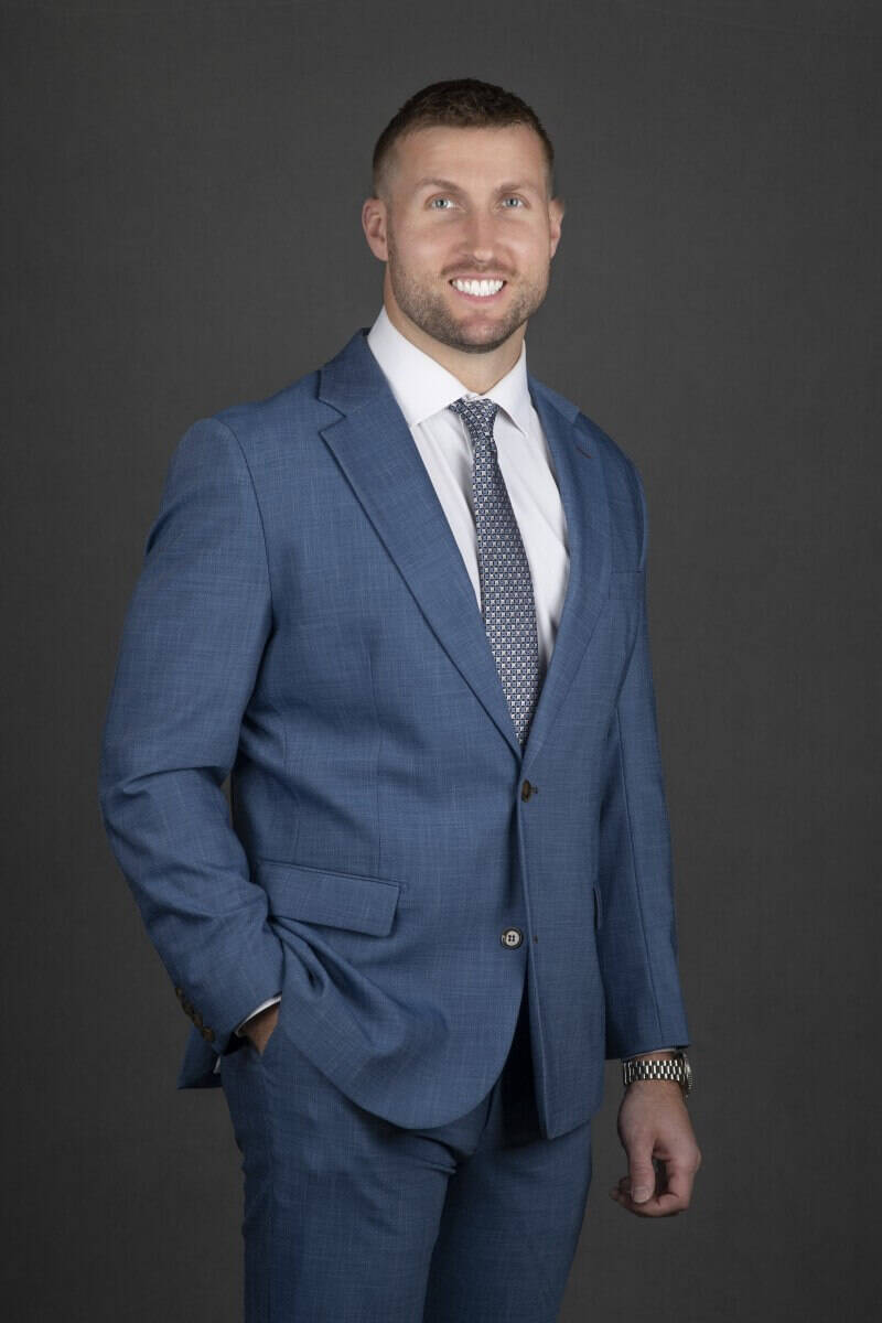 Professional man in a blue textured suit with a patterned tie and white dress shirt posing for a corporate headshot against a dark gray background