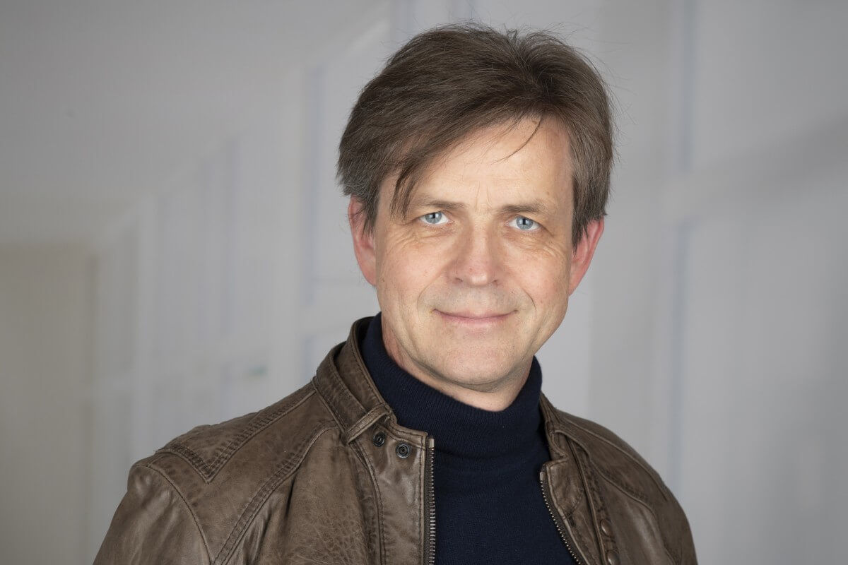 Portrait of a man with brown hair and blue eyes wearing a brown leather jacket and a navy blue turtleneck sweater against a soft, blurred background